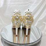 A pair of high heeled ivory satin shoes with a peep toe and a hidden platform at the front of the shoes and a gold crystal design on the back heel of the shoes