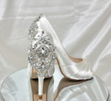 A pair of high heeled ivory satin shoes with a peep toe and a hidden platform at the front of the shoes and a crystal design on the back heel of the shoes