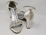 A pair of ivory block heel shoes with an ankle strap and a crystal design on the front toe strap of the shoes and a crystal design on the heel of the shoes
