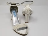 A pair of ivory block heel shoes with an ankle strap and a lace design on the front toe strap of the shoes and a lace design on the heel of the shoes