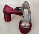 A pair of burgundy shoes with a low block heel and a peep toe front with a crystal design on the front of the shoes