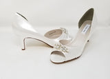 A pair of white satin bridal shoes with a peep toe and designed with a crystal design on the front of the shoes