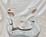 A pair of white block heel shoes with an ankle strap and a crystal design on the front toe strap of the shoes