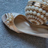 A pair of  bridal shoes in ivory satin with a kitten heel and a peep toe and a gold pearl and crystal design on the front of the shoes