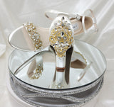 A pair of ivory block heel shoes with an ankle strap and a gold crystal design on the front toe strap of the shoes and a gold crystal design on the heel of the shoes