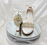 A pair of ivory block heel shoes with an ankle strap and a gold crystal design on the front toe strap of the shoes and a gold crystal design on the heel of the shoes