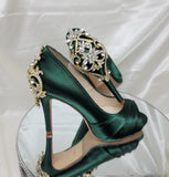 A pair of high heeled hunter green satin shoes with a peep toe and a hidden platform at the front of the shoes and a gold crystal design on the back heel of the shoes