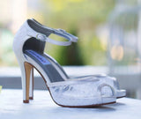 A pair of white lace high heeled shoes with a front hidden platform and an ankle strap with a peep toe