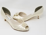 A pair of  bridal shoes in ivory satin with a kitten heel and a peep toe and a pearl and crystal design on the front of the shoes