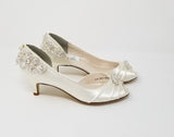A pair of  bridal shoes in ivory satin with a kitten heel and a peep toe and a crystal design on the front and back heel of the shoes