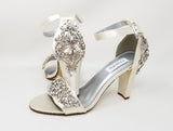 A pair of ivory high block heel shoes with an ankle strap and a crystal and pearl design on the front toe strap of the shoes and a large crystal design on the back heel of the shoes