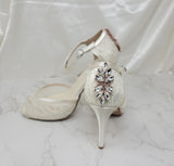 A pair of ivory high heeled platform lace shoes with an ankle strap and a crystal design on the back heel of the peep toe shoes