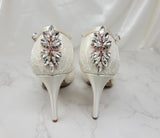 A pair of ivory high heeled platform lace shoes with an ankle strap and a crystal design on the back heel of the peep toe shoes