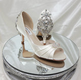 A pair of ivory satin medium height heel bridal shoes with a peep toe and designed with a crystal design on the front of the shoes and a crystal design on the back heel of the shoes
