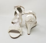 A pair of ivory wedding shoes with high wedge and straps across the front of the foot designed with a lace design on the front toe strap and a lace design on the back heel of the shoes