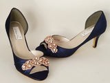 A pair of navy blue satin medium height heel shoes with a peep toe and designed with a rose gold crystal design on the front of the shoes