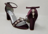 A pair of eggplant purple block heel shoes with an ankle strap and a crystal design on the front toe strap of the shoes and a crystal design on the heel of the shoes