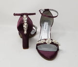 A pair of eggplant purple block heel shoes with an ankle strap and a crystal and pearl design on the front toe strap of the shoes and a crystal and pearl design on the heel of the shoes
