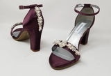 A pair of eggplant purple block heel shoes with an ankle strap and a crystal and pearl design on the front toe strap of the shoes and a crystal and pearl design on the heel of the shoes