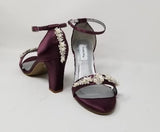 A pair of eggplant purple block heel shoes with an ankle strap and a crystal and pearl design on the front toe strap of the shoes and a crystal and pearl design on the heel of the shoes