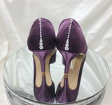 A pair of eggplant purple satin medium height heel bridal shoes with a peep toe and designed with a crystal design on the front of the shoes and a crystal teardrop design on the back heel of the shoes