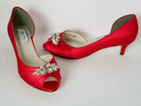 A pair of red satin kitten heel shoes with a peep toe and a rose gold crystal design on the front of the shoes