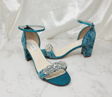 A pair of green lace covered low block heel shoes with an ankle strap and a crystal design on the front toe strap of the shoes