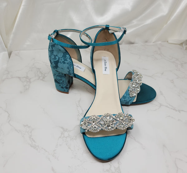 A pair of green lace covered low block heel shoes with an ankle strap and a crystal design on the front toe strap of the shoes