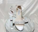 A pair of white block heel shoes with an ankle strap and a crystal design on the front toe strap of the shoes