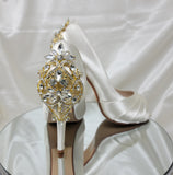 A pair of high heeled ivory satin shoes with a peep toe and a hidden platform at the front of the shoes and a gold crystal design on the back heel of the shoes