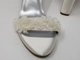 A pair of ivory block heel shoes with an ankle strap and a lace design on the front toe strap of the shoes and a lace design on the heel of the shoes