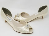 A pair of ivory satin kitten heel shoes with a peep toe with a small crystal design on the front of the shoes