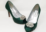 A pair of high heeled hunter green satin shoes with a peep toe and a hidden platform at the front of the shoes and a crystal design on the front of the shoes