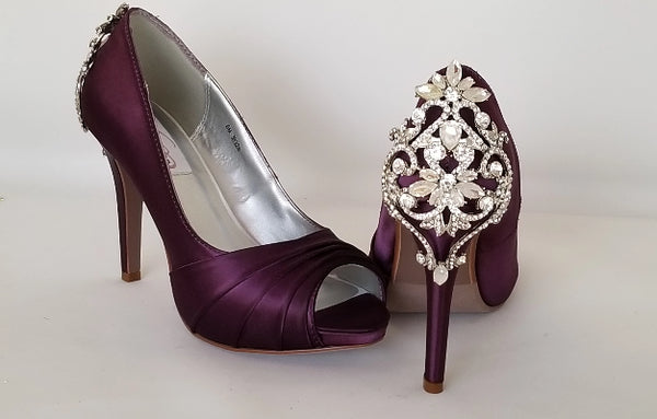 A pair of high heeled eggplant  purple satin shoes with a peep toe and a hidden platform at the front of the shoes and a crystal design on the back heel of the shoes