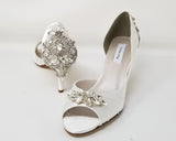 A pair of white satin bridal shoes with a peep toe and designed with a crystal design on the front of the shoes and a large crystal design on the back heel of the shoes