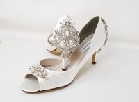 A pair of white satin bridal shoes with a peep toe and designed with a crystal design on the front of the shoes and a large crystal design on the back heel of the shoes