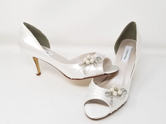 A pair of white satin bridal shoes with a peep toe and designed with a pearl and crystal bow design on the side of the shoes