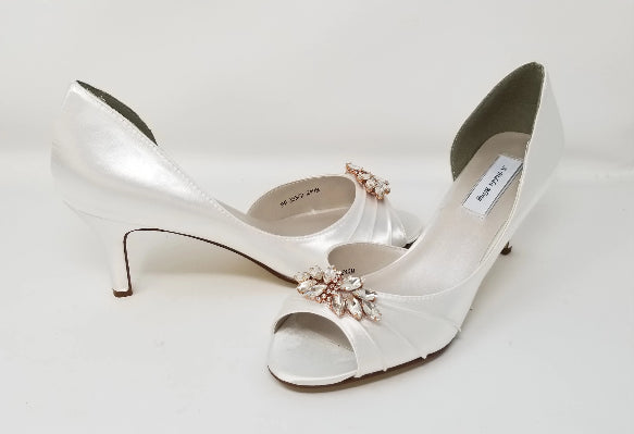 A pair of white satin bridal  heels with a peep toe and designed with a rose gold crystal design on the front of the shoes 