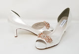 A pair of white satin bridal shoes with a peep toe and designed with a rose gold crystal design on the front of the shoes
