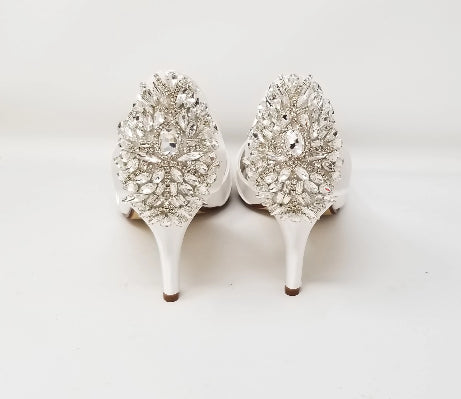 A pair of white satin bridal shoes with a peep toe and designed with a crystal design on the front of the shoes and a large crystal design on the back heel of the shoes