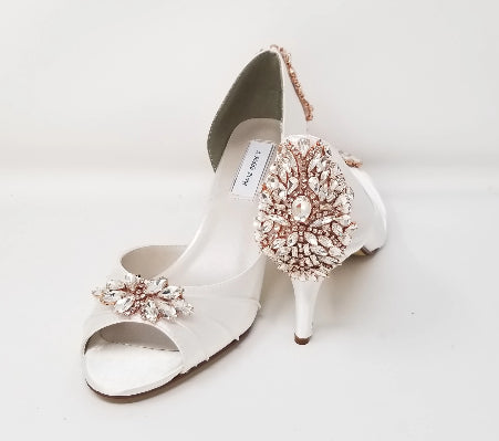 A pair of white satin heels with a peep toe and designed with a rose gold crystal design on the front of the shoes and a large rose gold crystal design on the back heel of the shoes