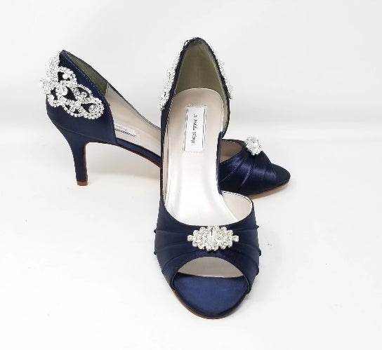 A pair of navy blue satin medium height heel shoes with a peep toe and designed with a crystal design on the front of the shoes shoes and a crystal design on the back heel of the shoes