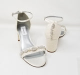 A pair of ivory lace covered low block heel shoes with an ankle strap and a crystal design on the front toe strap of the shoes and a crystal design on the back heel o the shoes