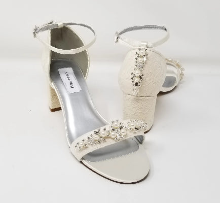 A pair of ivory lace covered low block heel shoes with an ankle strap and a crystal and pearl design on the front toe strap of the shoes and a crystal and pearl design on the back heel of the shoes