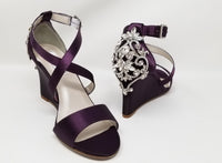 A pair of eggplant purple wedding shoes with high wedge and straps across the front of the foot designed with a crystal design on the front toe strap of the shoes and a crystal design on the back heel of the shoes