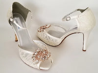 A pair of ivory high heeled platform lace shoes with an ankle strap and a rose gold crystal design on the front of the peep toe shoes