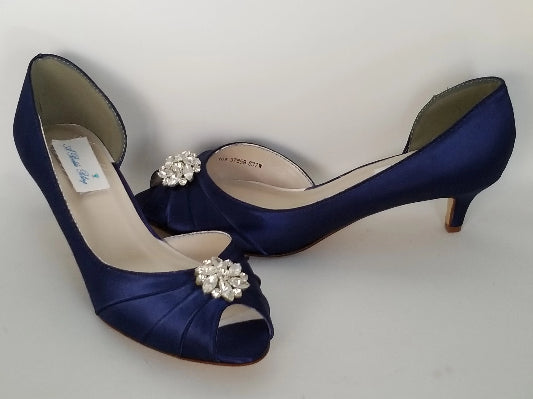 A pair of  bridal shoes in navy blue satin with a kitten heel and a peep toe and a crystal design on the front of the shoes