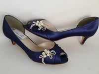 A pair of  bridal shoes in navy blue satin with a kitten heel and a peep toe and a pearl and crystal bow design on the side of the shoes