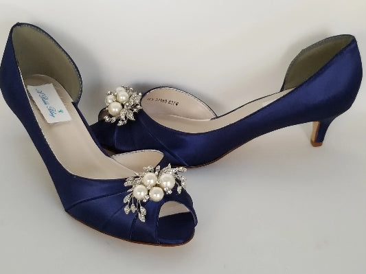 A pair of  bridal shoes in navy blue satin with a kitten heel and a peep toe and a pearl and crystal design on the front of the shoes
