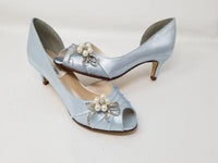 A pair of baby blue satin kitten heels with a peep toe and designed with a pearl and crystal bow design on the side of the shoes